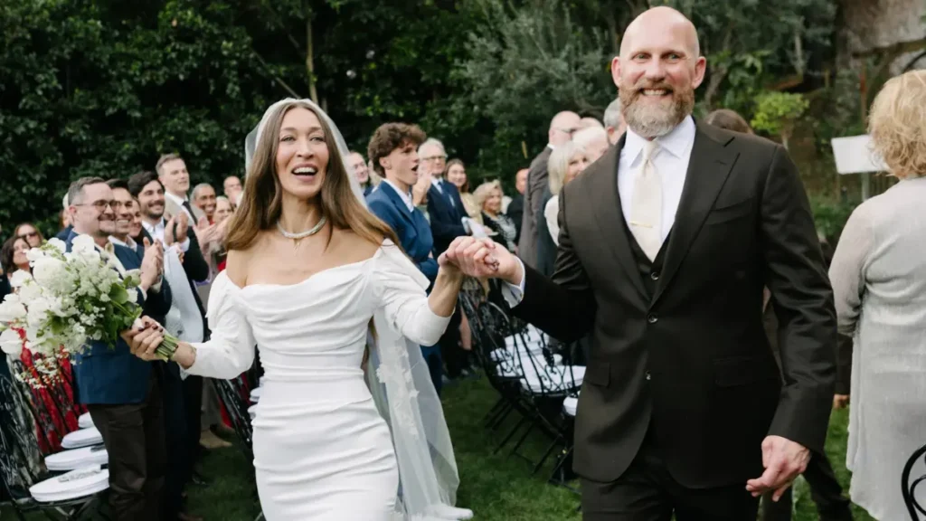 Alana Hadid standing hand-in-hand with her partner, outdoors in daylight on the grounds of her father’s Los Angeles home, she wearing a vintage off-white gown and he in a dark suit, surrounded by family and greenery.