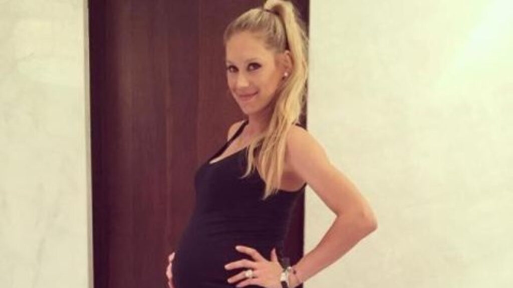 Anna Kournikova pregnant in 2025, smiling and cradling her baby bump at home.