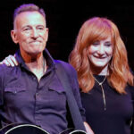 Bruce Springsteen and Patti Scialfa smiling together at an event, exuding warmth and love as they stand side by side, with Bruce in a black jacket and Patti in a stylish outfit.