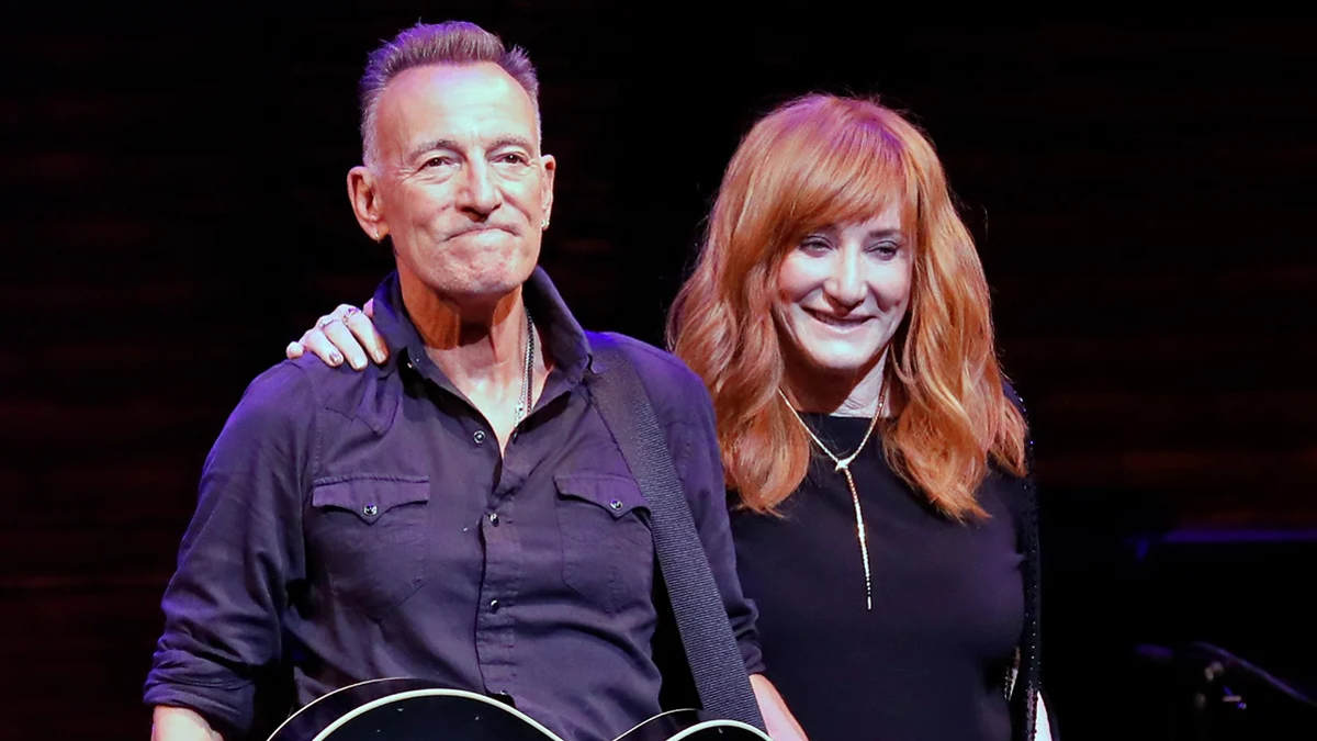 Bruce Springsteen and Patti Scialfa smiling together at an event, exuding warmth and love as they stand side by side, with Bruce in a black jacket and Patti in a stylish outfit.