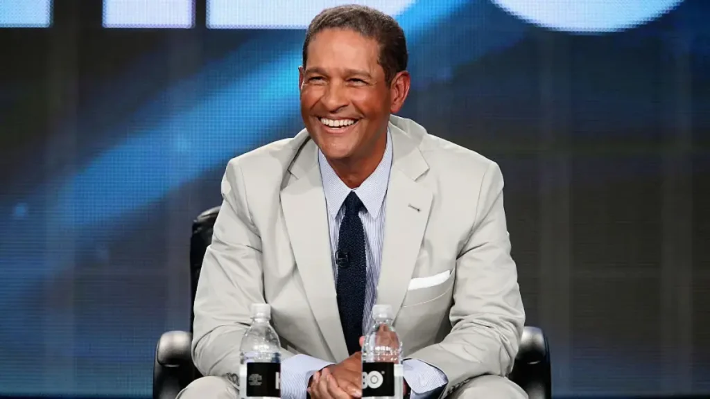 Bryant Gumbel seated on a studio chair in a white suit, legs crossed, speaking during an interview segment.