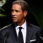 Bryant Gumbel wearing a dark suit and patterned tie, standing at a podium on stage.