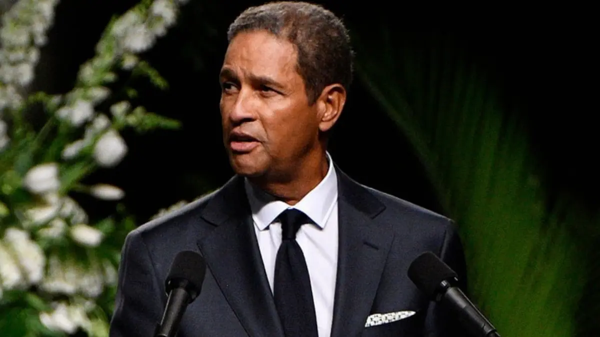 Bryant Gumbel wearing a dark suit and patterned tie, standing at a podium on stage.