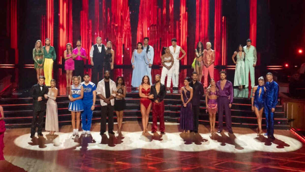 All Dancing With the Stars contestants standing side by side on stage under bright lights, waiting tensely as the elimination results are announced.