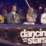 Panel of judges on stage: female judge in an elegant sheer-sleeved dress, male judge in crisp white tuxedo, another male judge wearing a metallic gold blazer. The ‘Dancing With the Stars’ sign glows on the front of their desk.