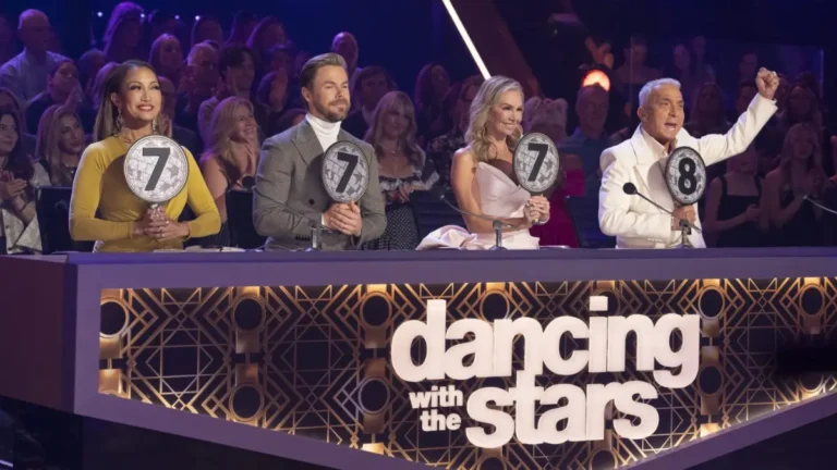 Panel of judges on stage: female judge in an elegant sheer-sleeved dress, male judge in crisp white tuxedo, another male judge wearing a metallic gold blazer. The ‘Dancing With the Stars’ sign glows on the front of their desk.