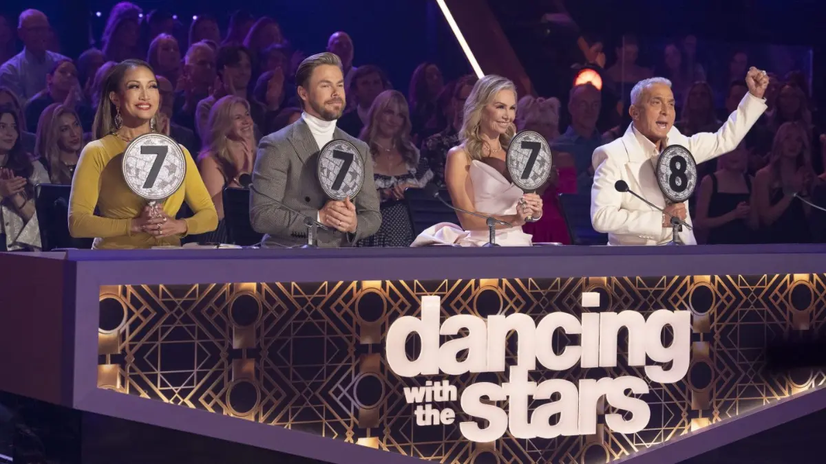 Panel of judges on stage: female judge in an elegant sheer-sleeved dress, male judge in crisp white tuxedo, another male judge wearing a metallic gold blazer. The ‘Dancing With the Stars’ sign glows on the front of their desk.