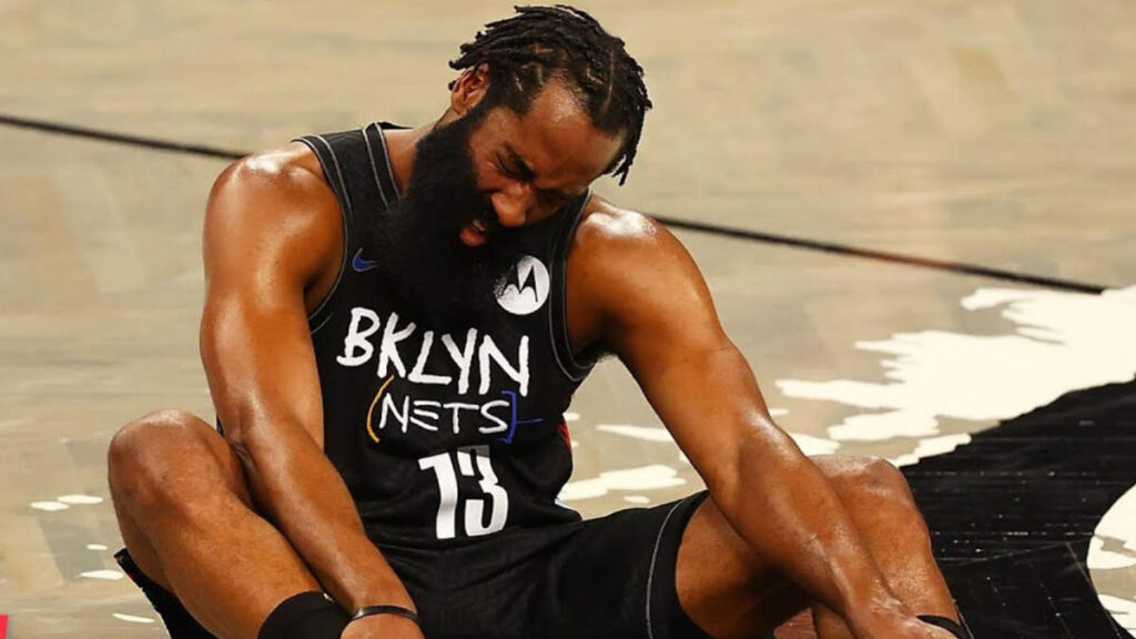 James Harden grimacing as he clutches his hamstring mid-play on the court, referee signaling timeout.