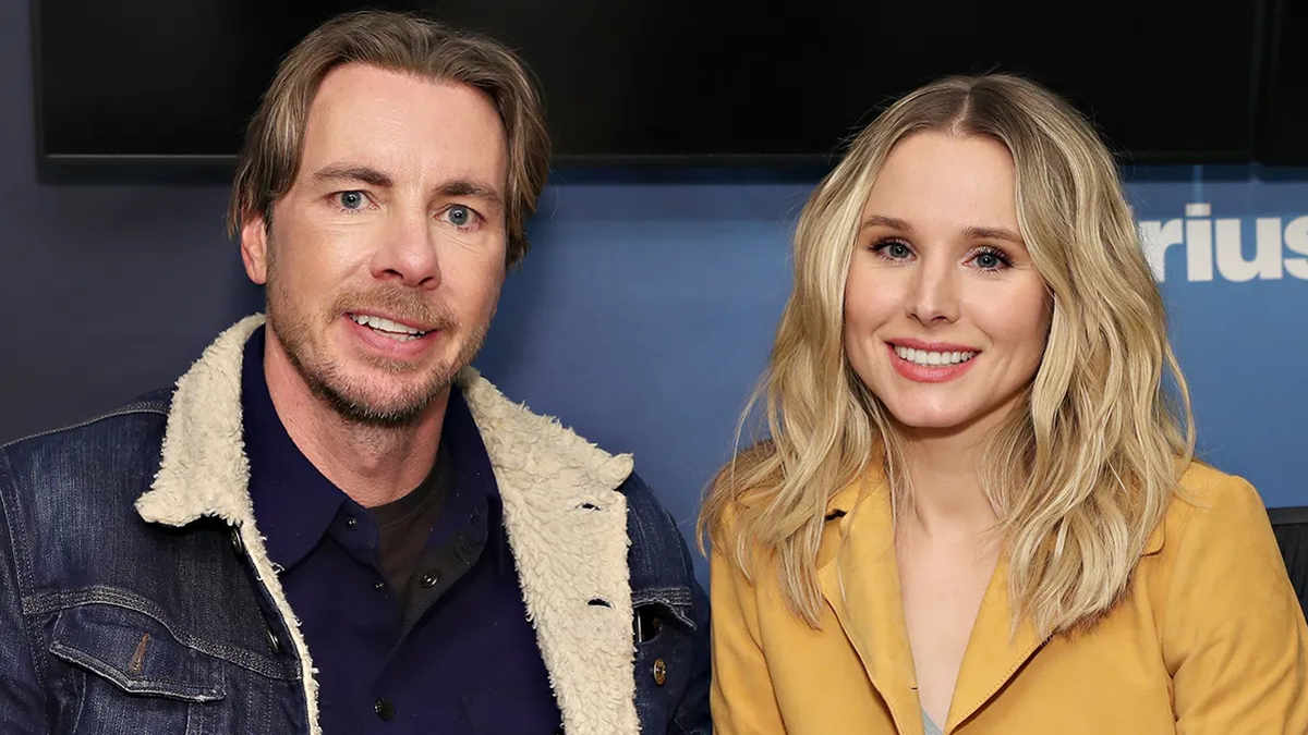 Kristen Bell and Dax Shepard standing together on set, smiling at the camera, promoting their latest project.