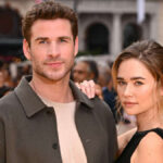 Liam Hemsworth and Gabriella Brooks posing together on the red carpet, both smiling, dressed in formal attire, with cameras flashing in the background.