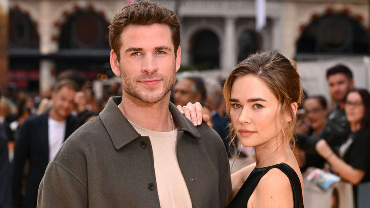 Liam Hemsworth and Gabriella Brooks posing together on the red carpet, both smiling, dressed in formal attire, with cameras flashing in the background.