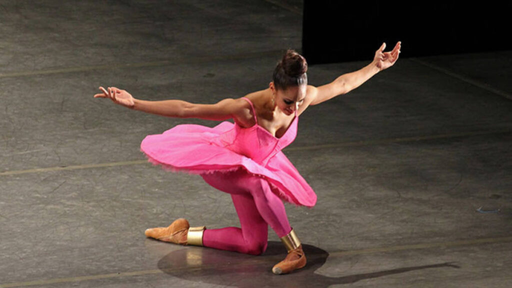 Misty Copeland standing center stage in a red costume, one arm raised elegantly as the spotlight shines down on her.