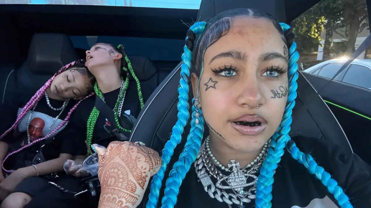 North West displaying playful tattoo designs on her face — a small star under her right eye and her name in cursive script on her left cheek, paired with bold braids and colored contact lenses.
