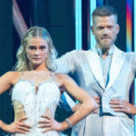 Scott Hoying and Rylee Arnold standing side by side on stage, holding hands as the host announces their elimination, stage lights glowing softly behind them.