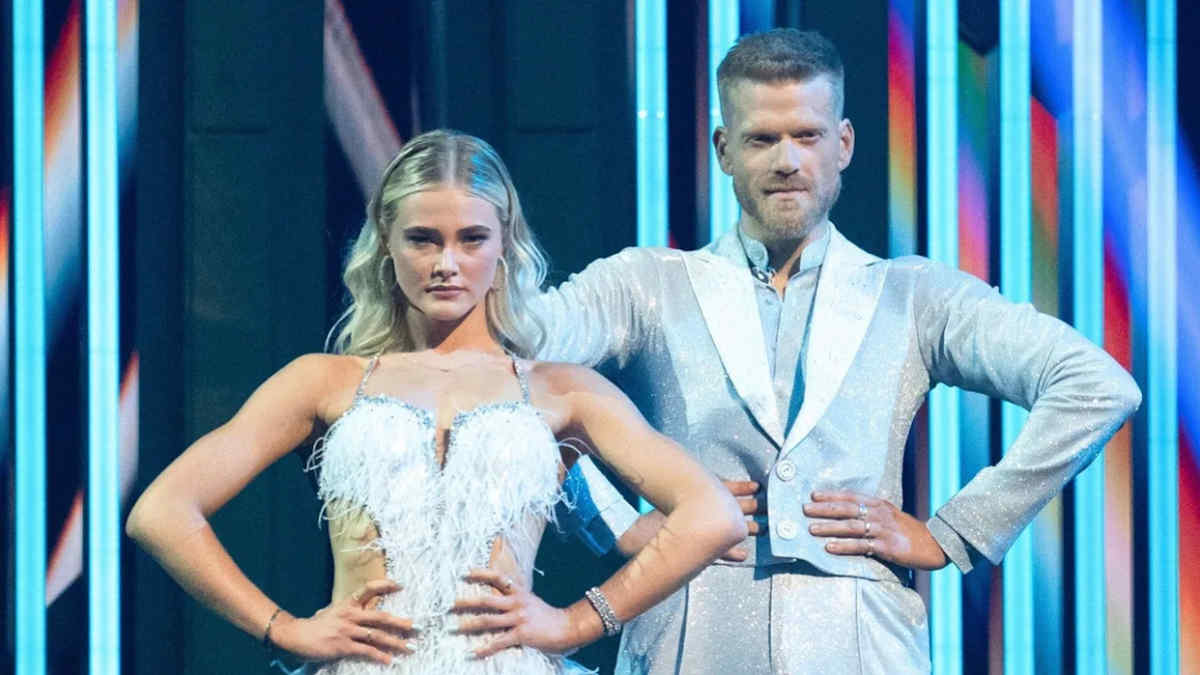 Scott Hoying and Rylee Arnold standing side by side on stage, holding hands as the host announces their elimination, stage lights glowing softly behind them.