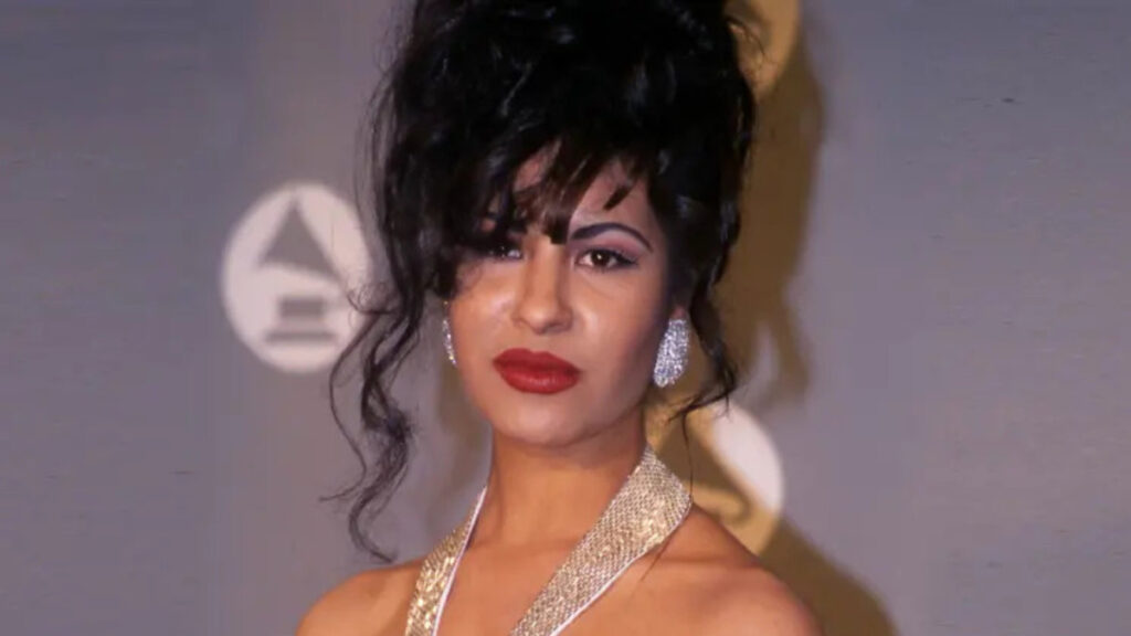 Close-up portrait of Selena Quintanilla with dark wavy hair, red lipstick, and gold hoop earrings, softly smiling against a neutral background.