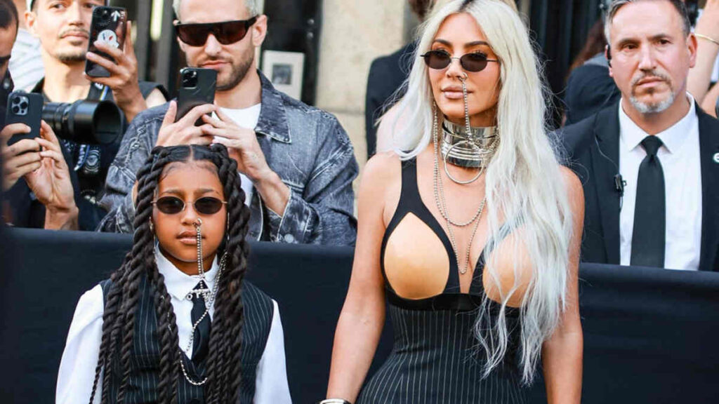 Kim Kardashian and her daughter North West standing side by side at a public event, both leaning slightly forward and peering toward a loud noise off-camera.